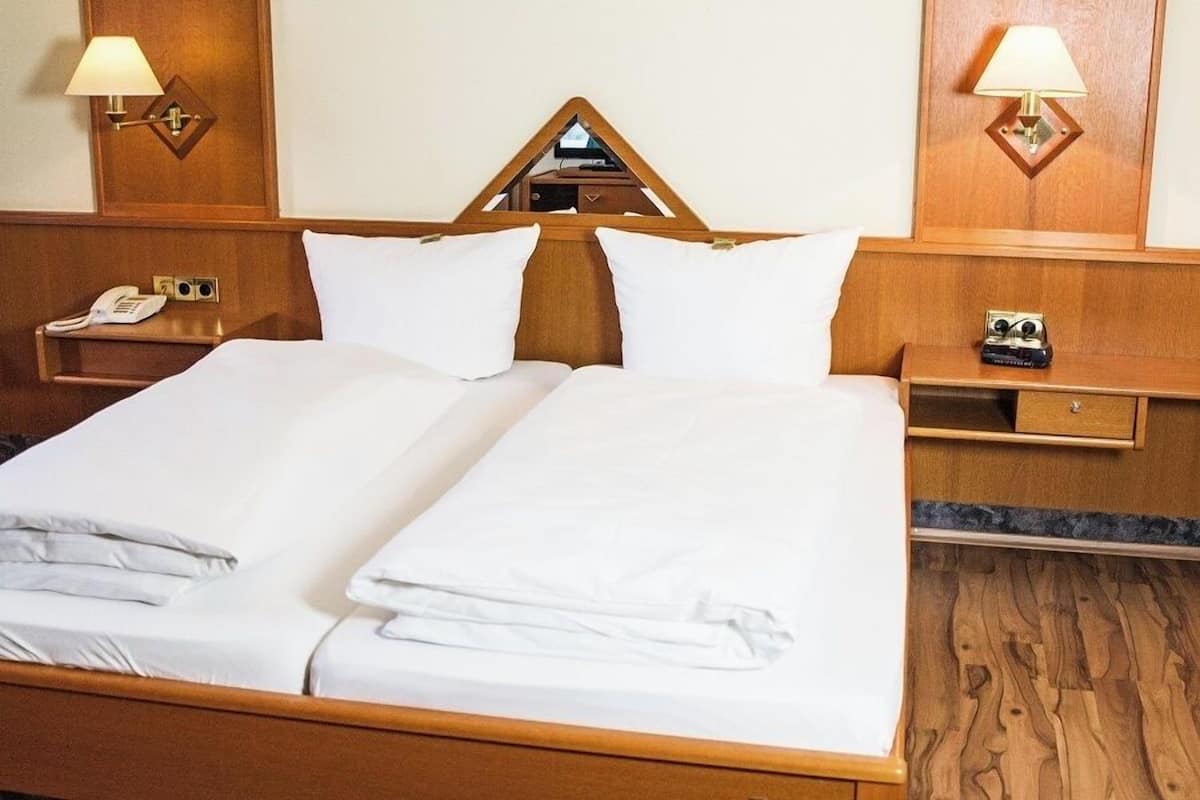 Un lit bien fait, avec du linge de lit blanc, une tête de lit en bois et une table de chevet sur laquelle se trouvent une lampe et un téléphone.