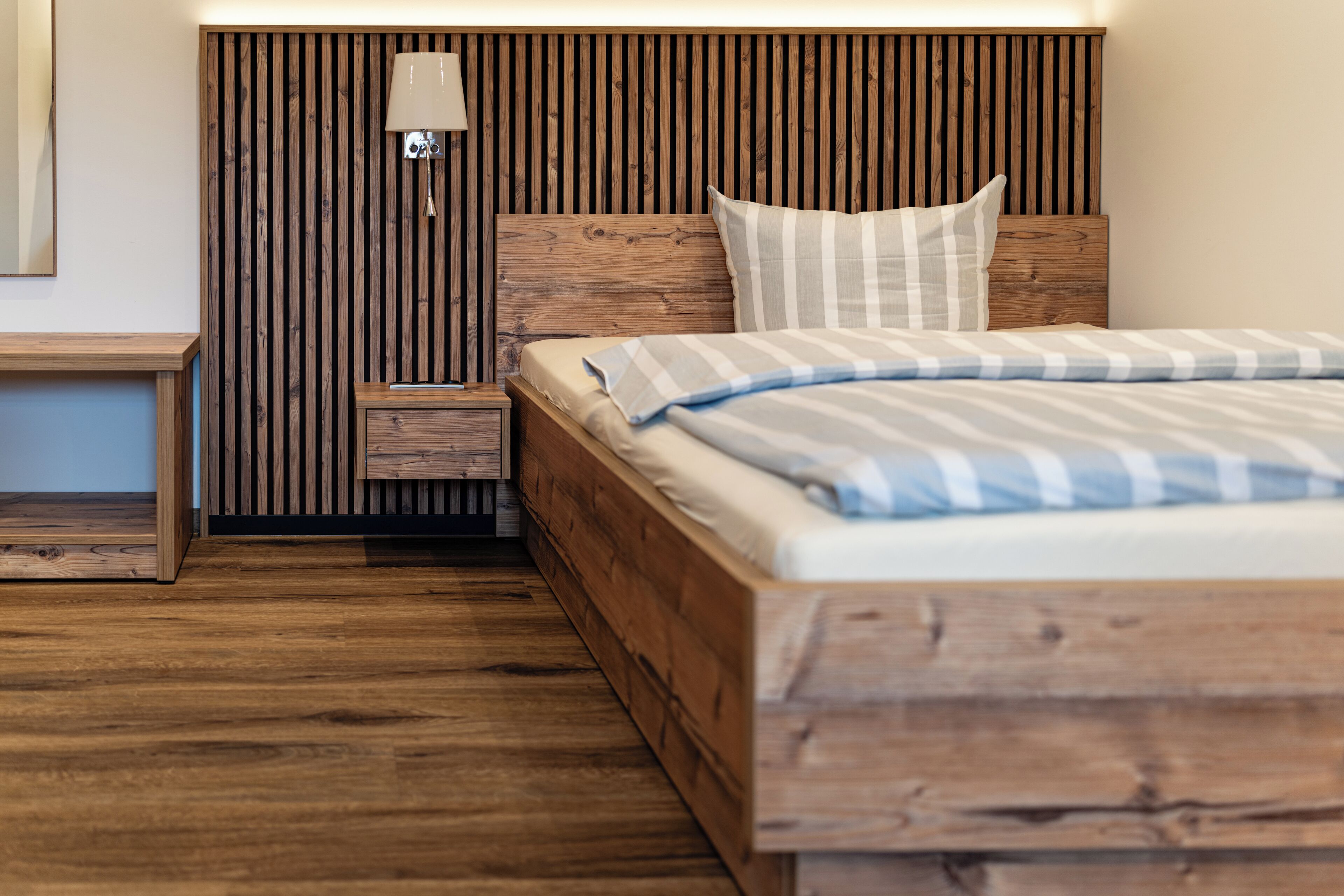 Ein hölzernes Bett mit gestreifter Decke, ein Nachttisch mit Lampe und ein Holzkopfende mit vertikalen Lamellen.