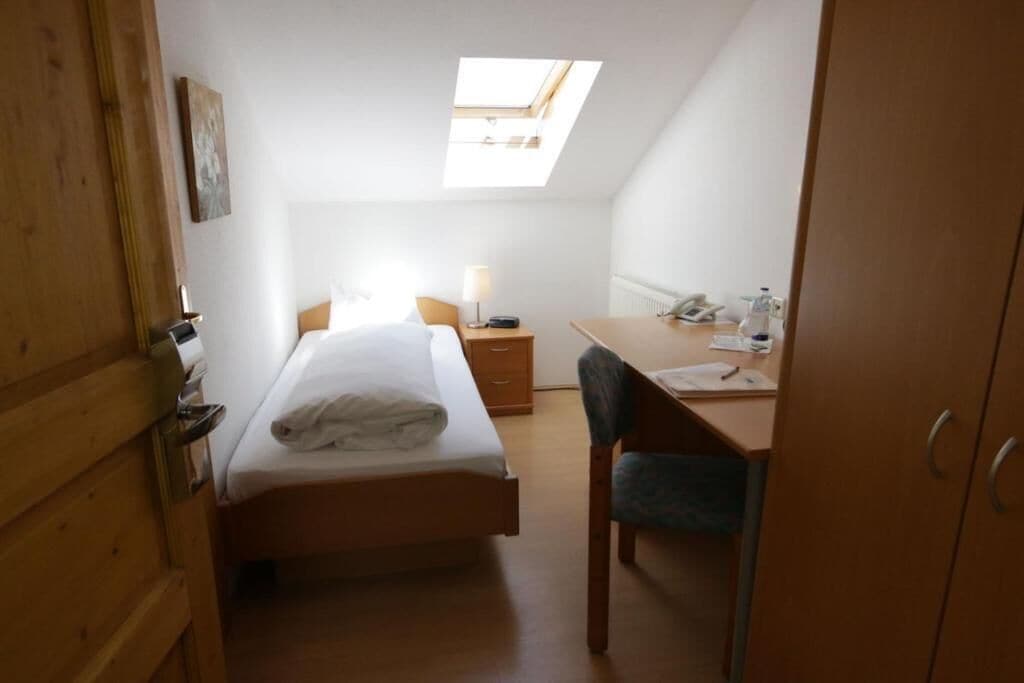 Ein kleiner, kompakter Raum mit einem Bett, einem Schreibtisch und einem Stuhl. Über dem Bett befindet sich ein Oberlicht, und links eine Holztür.