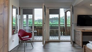 Una habitación moderna con piso de madera, una silla roja, un televisor y un balcón con terraza de madera y vegetación.