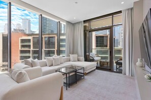 A modern living room with a large sofa, a coffee table, and a balcony with a view of the cityscape. - Lavish 1-Bedroom w/ Burj Khalifa view in Peninsula (Business Bay)