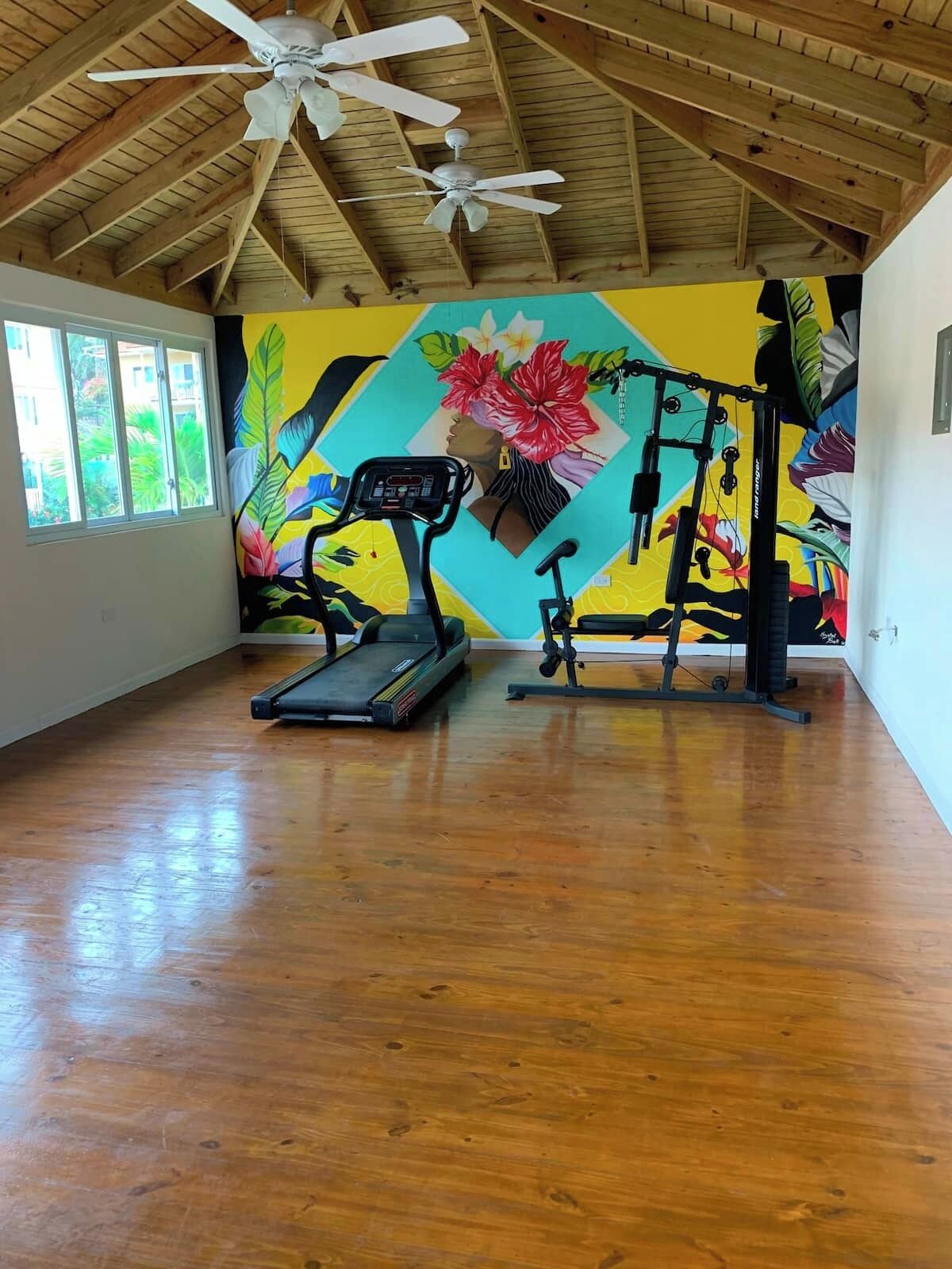 A gym with a treadmill, stationary bike, and weight machine, featuring a vibrant mural on the wall.