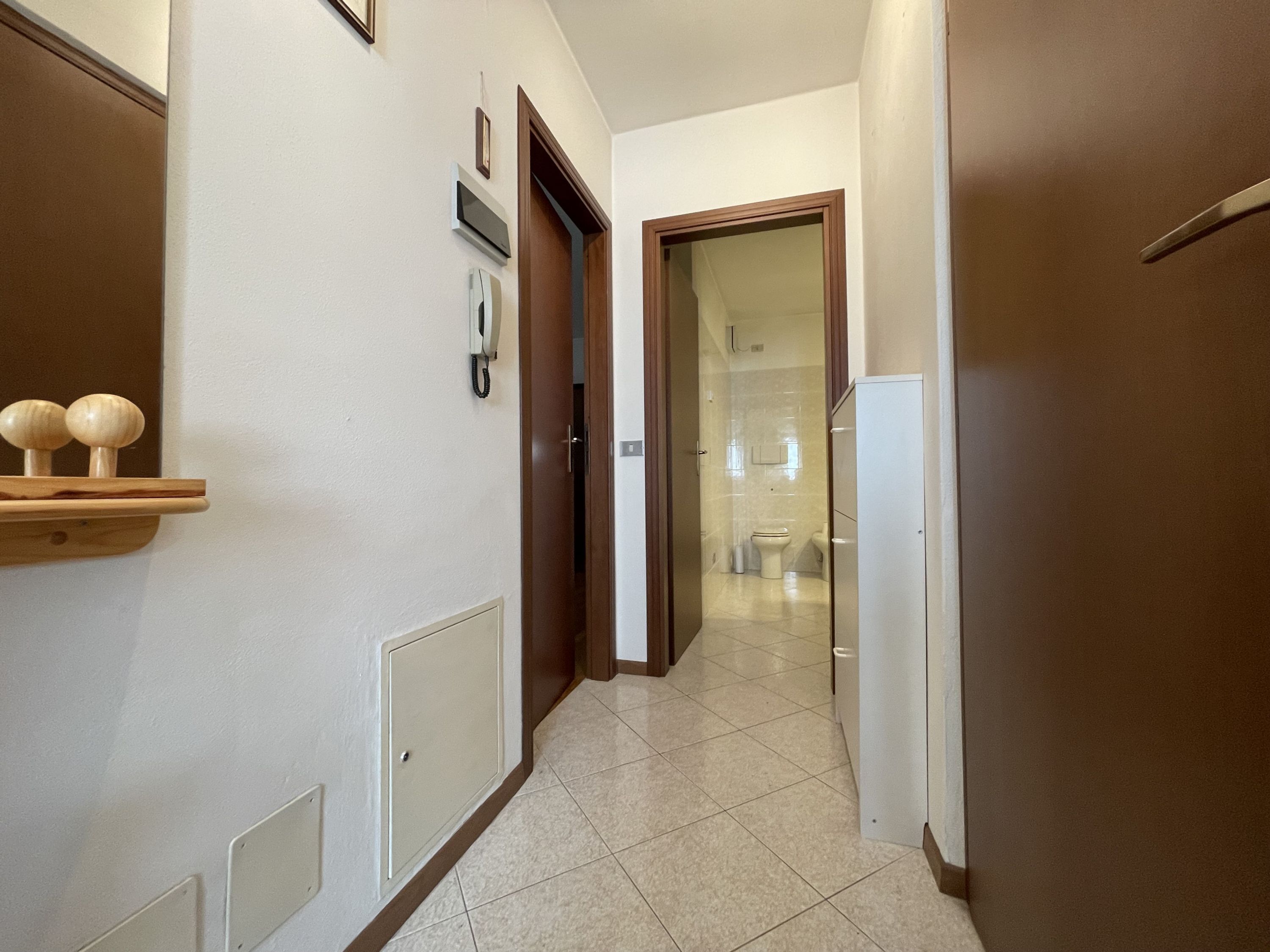 A hallway with a wooden door, a white wall with a phone, and a tiled floor.