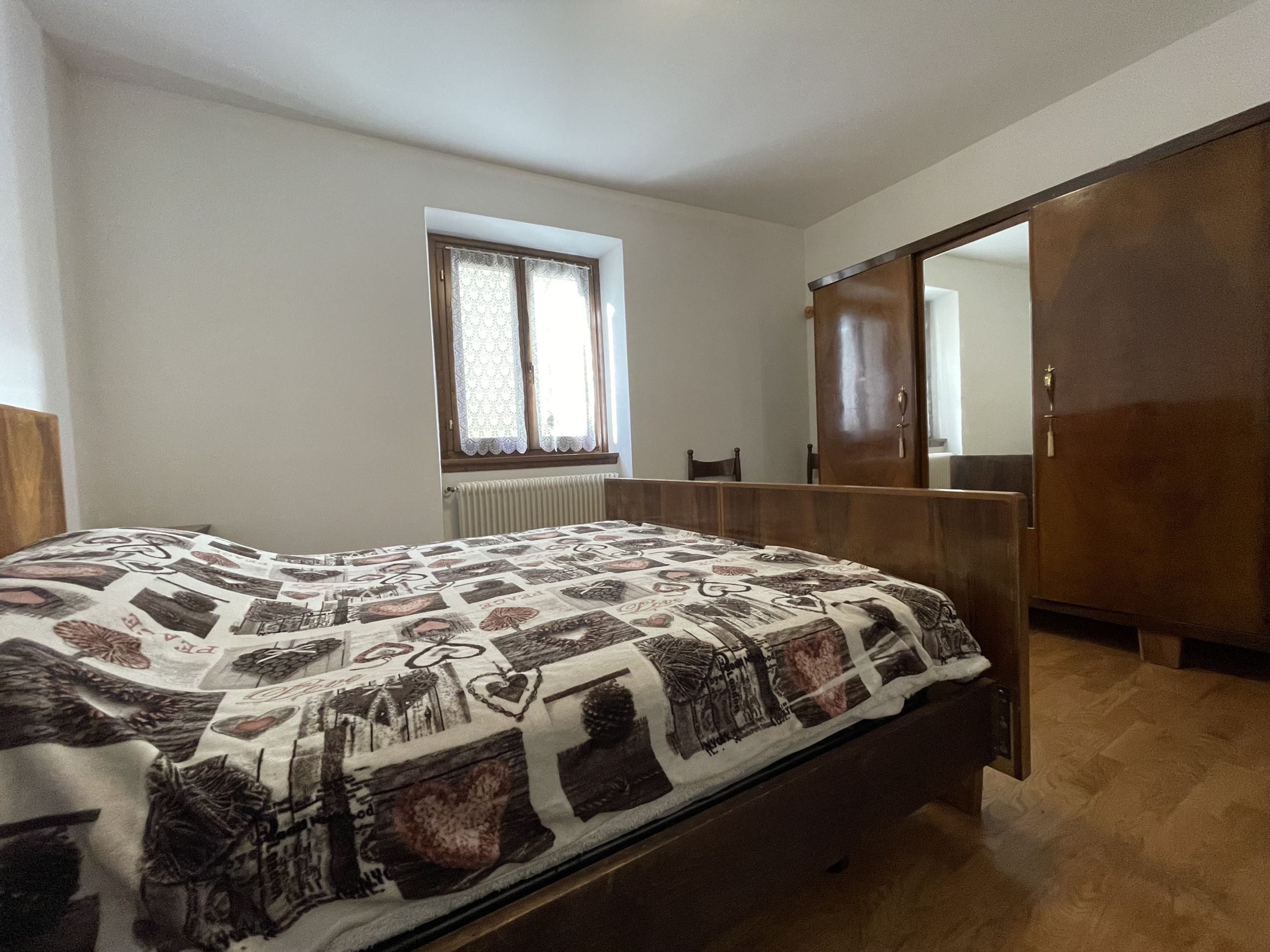 Ein Schlafzimmer mit Bett, einem Fenster mit Spitzenvorhängen, einer Heizung, einem Holzschrank und einem Spiegel an der Wand.
