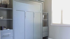 Un armario gris con dos puertas y una repisa, un escritorio pequeño con cajones, y una ventana con vistas.