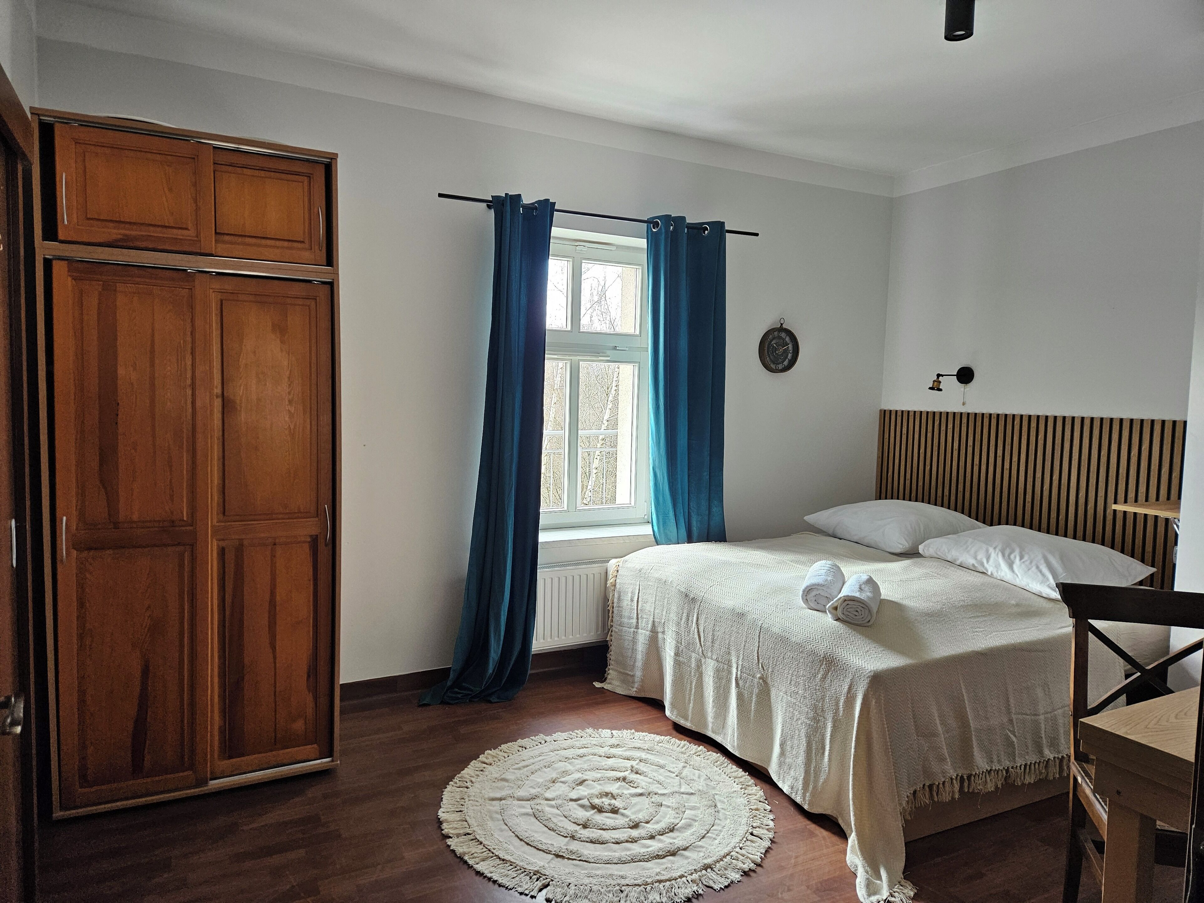 A bedroom with a bed, a wooden wardrobe, a window with curtains, a bedside table, and a wall clock.