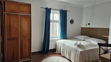 A bedroom with a bed, a wooden wardrobe, a window with curtains, a bedside table, and a wall clock.