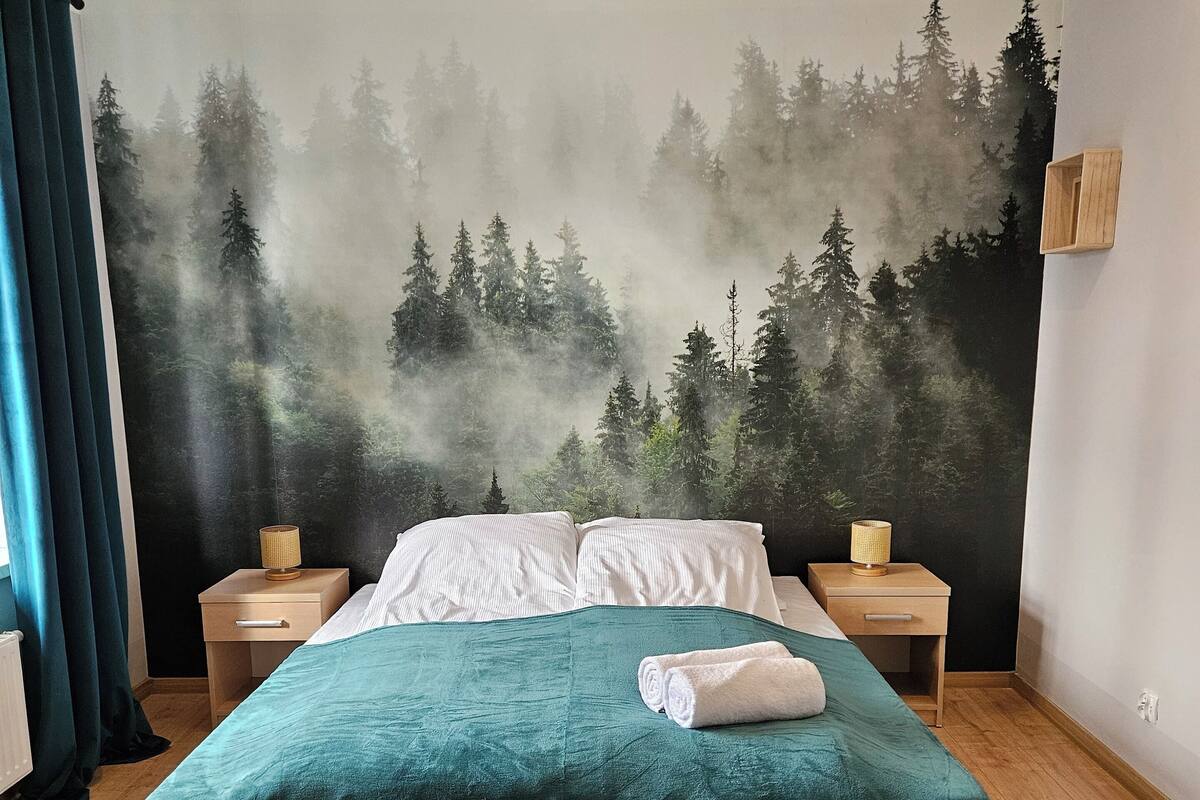 A bedroom with a bed, two bedside tables, a wall mural, and a round rug.