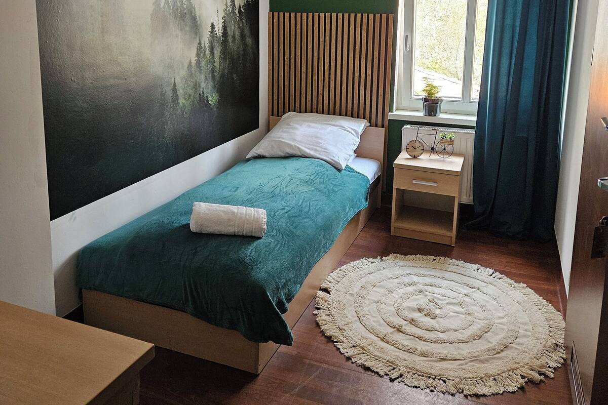 A bedroom with a bed, a nightstand, a window with curtains, a wall mural, and a round rug.