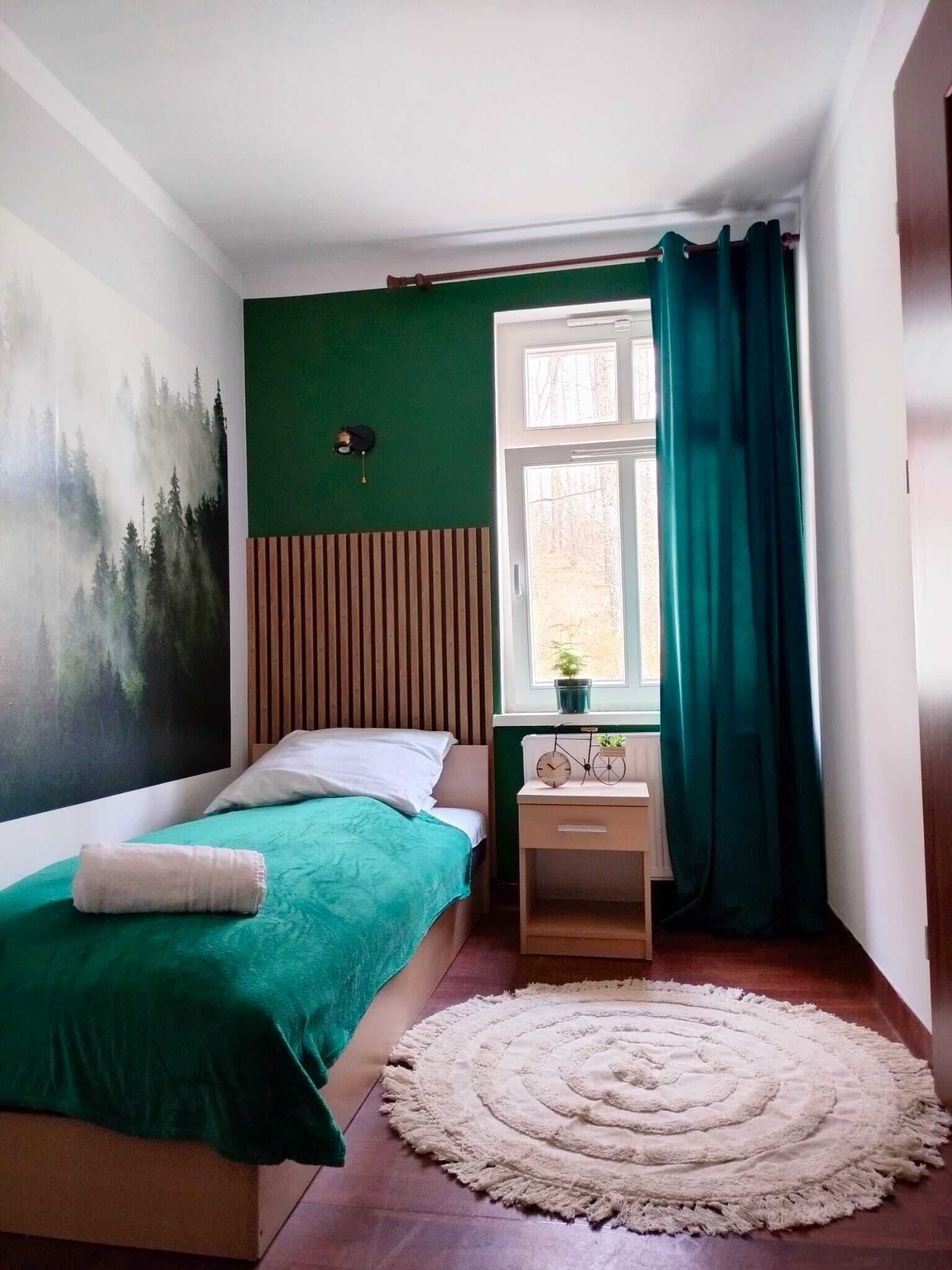 Un dormitorio con cama, mesita de noche, ventana con cortinas, un cuadro en la pared y alfombra.
