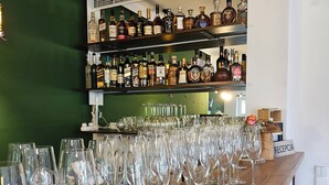 Una zona de bar con múltiples estantes llenos de botellas de licores y una fila de copas de vino vacías sobre una encimera de madera.
