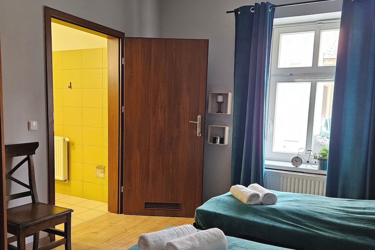 A room with two beds, a wooden chair, a radiator, and a window with curtains.