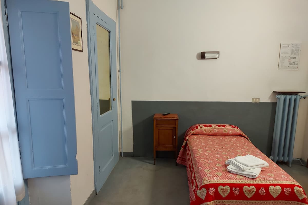 Une chambre avec un lit simple, une petite table de chevet en bois, une porte bleue et un interrupteur sur le mur.