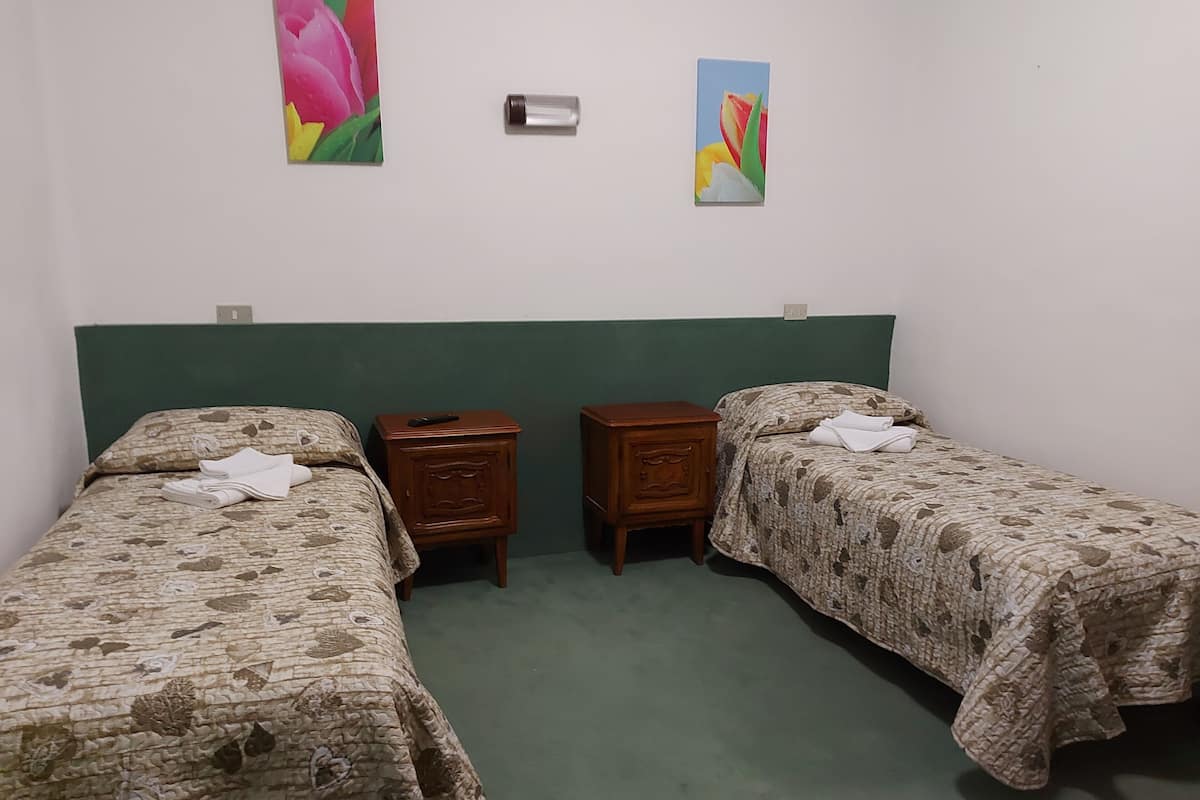 Une chambre avec deux lits simples, chacun recouvert d’une couvre-lit à motifs floraux, une table de chevet en bois avec une télécommande, et une tête de lit verte.