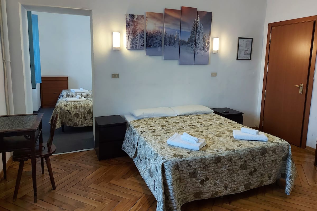 Une chambre d’hôtel avec un grand lit, un plus petit lit, un bureau et une chaise. Il y a des lampes fixées au mur et une porte.