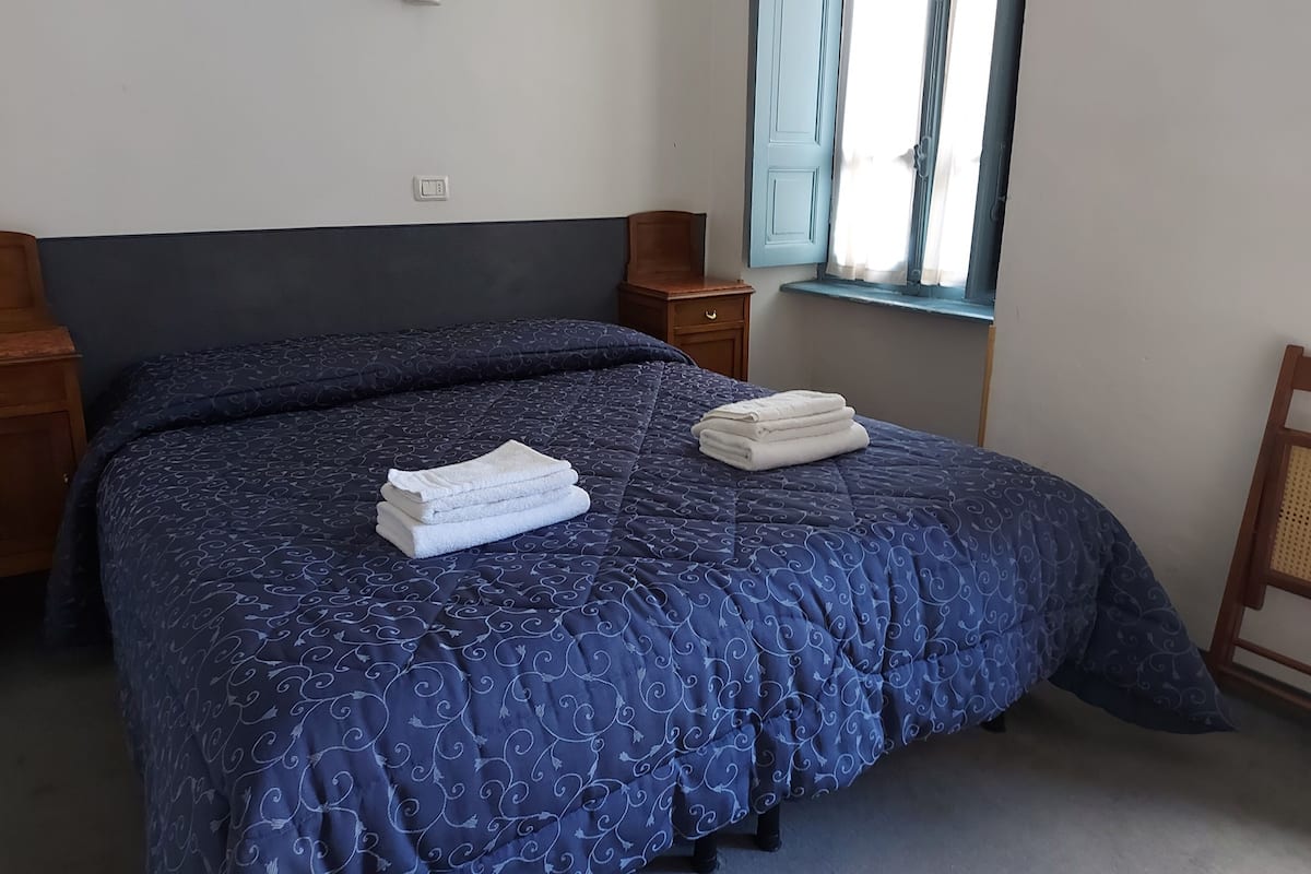 Un lit bien fait, recouvert d’une couvre-lit bleu foncé à motifs, deux serviettes blanches pliées, une table de chevet en bois avec une lampe, et une fenêtre avec des rideaux.