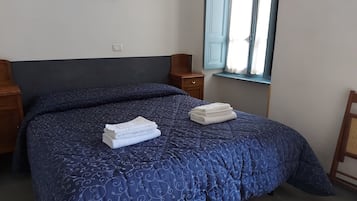A neatly made bed with a dark blue patterned bedspread, two folded white towels, a wooden nightstand with a lamp, and a window with curtains.