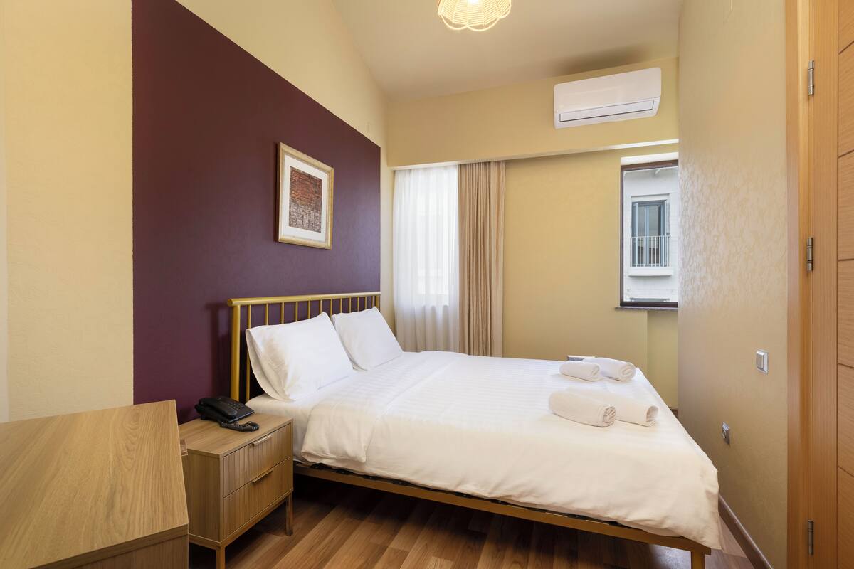 Quarto de hotel com cama de madeira, mesa de cabeceira com telefone, janela com cortinas e ar condicionado.