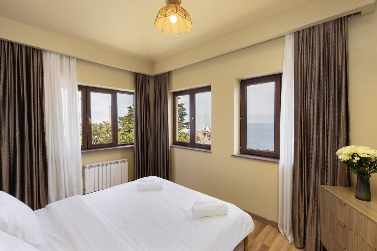 Quarto de hotel com duas camas, cómoda de madeira, um vaso de flores e amplas janelas com cortinas.