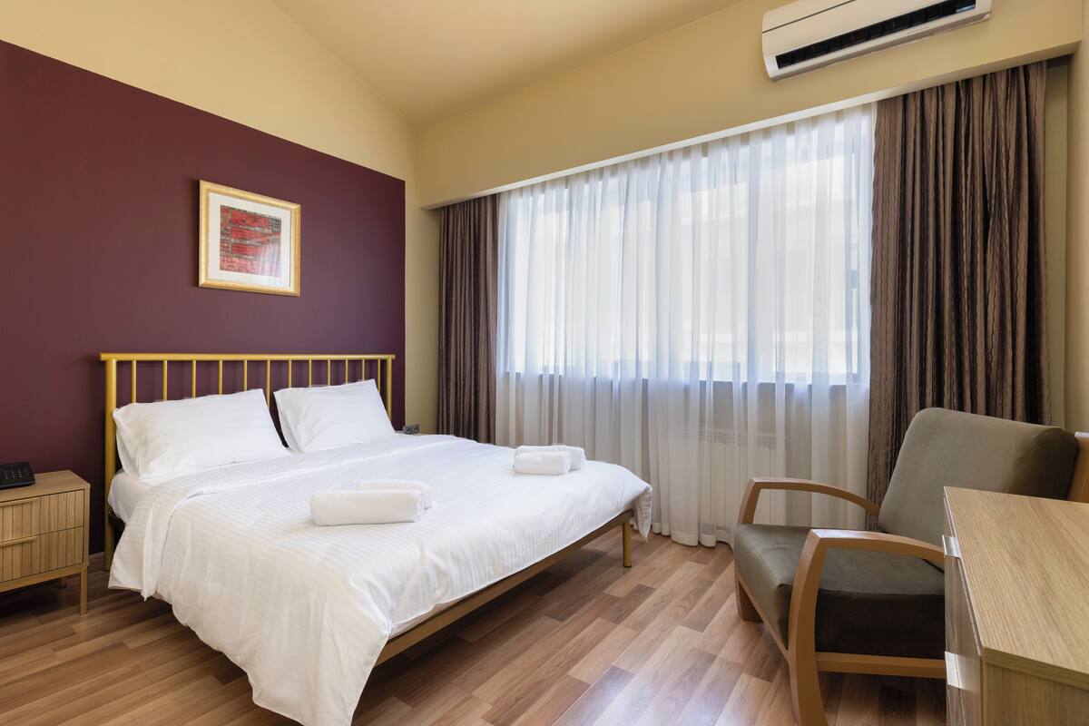 Quarto de hotel com cama, cadeira, mesa, mesa de cabeceira e janela com cortinas.