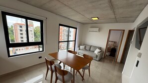 Amplia sala de estar con mesa y sillas de comedor, un sofá y ventanas grandes que ofrecen vistas de edificios.
