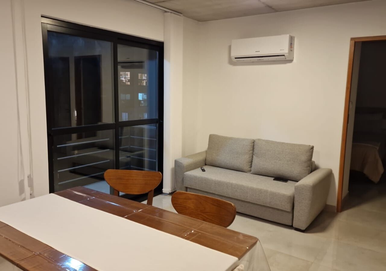 Una sala de estar con sofá, una mesa de comedor y sillas. Hay una puerta corrediza de vidrio que da a un balcón.