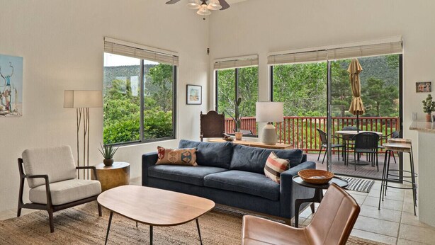 A spacious living room with a sofa, armchairs, and a coffee table. Large windows offer a view of greenery outside.