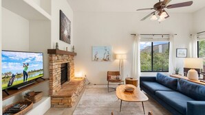 A living room with a fireplace, a television, a sofa, a ceiling fan, and a view of the outdoors.