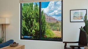 A room with a large window offering a view of a forest and mountains. There is a blue sofa, a wooden table with a plant, and a framed picture on the wall.