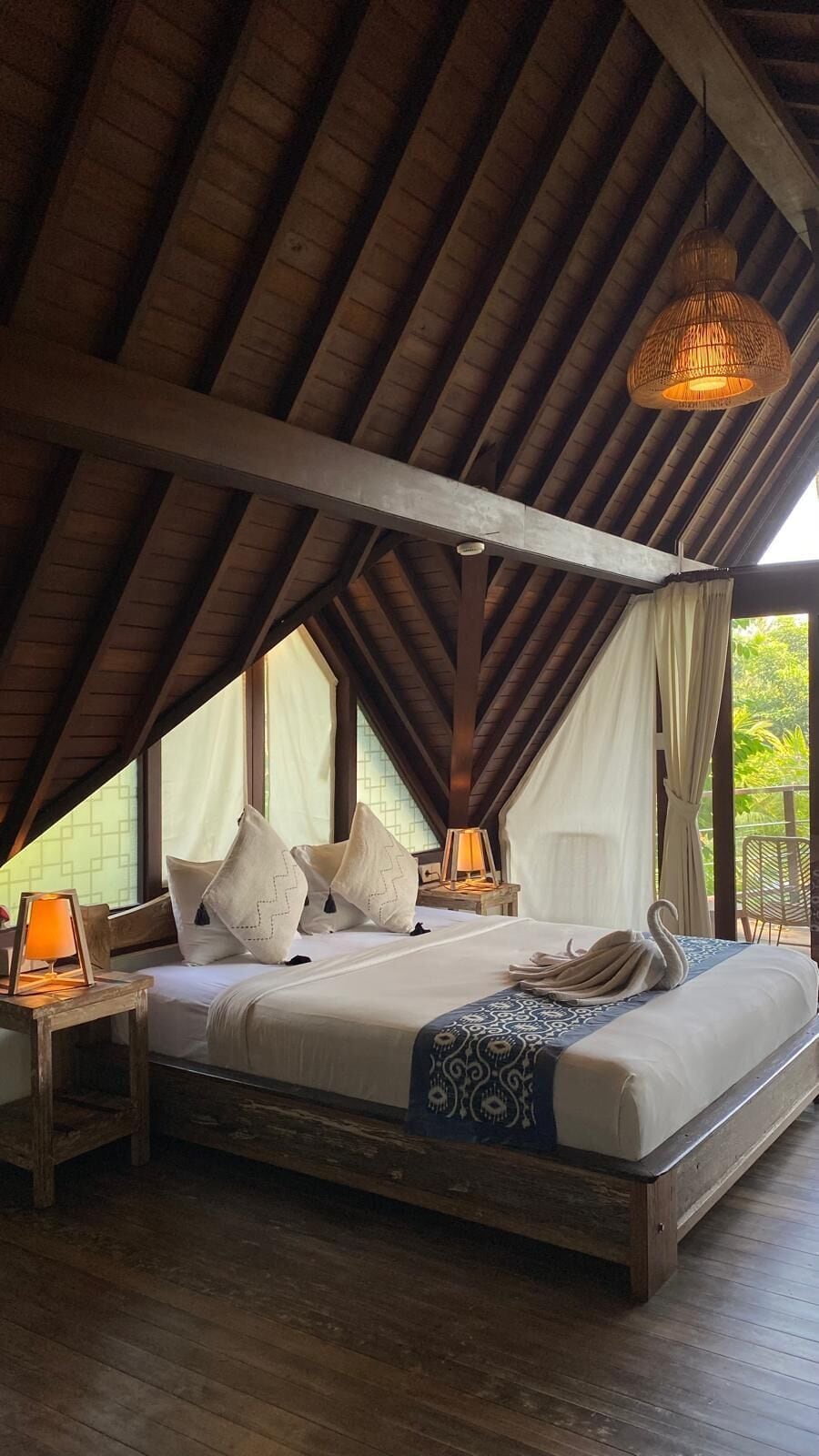 A bedroom with a wooden ceiling, a bed with pillows, a bedside lamp, and a hanging light fixture.