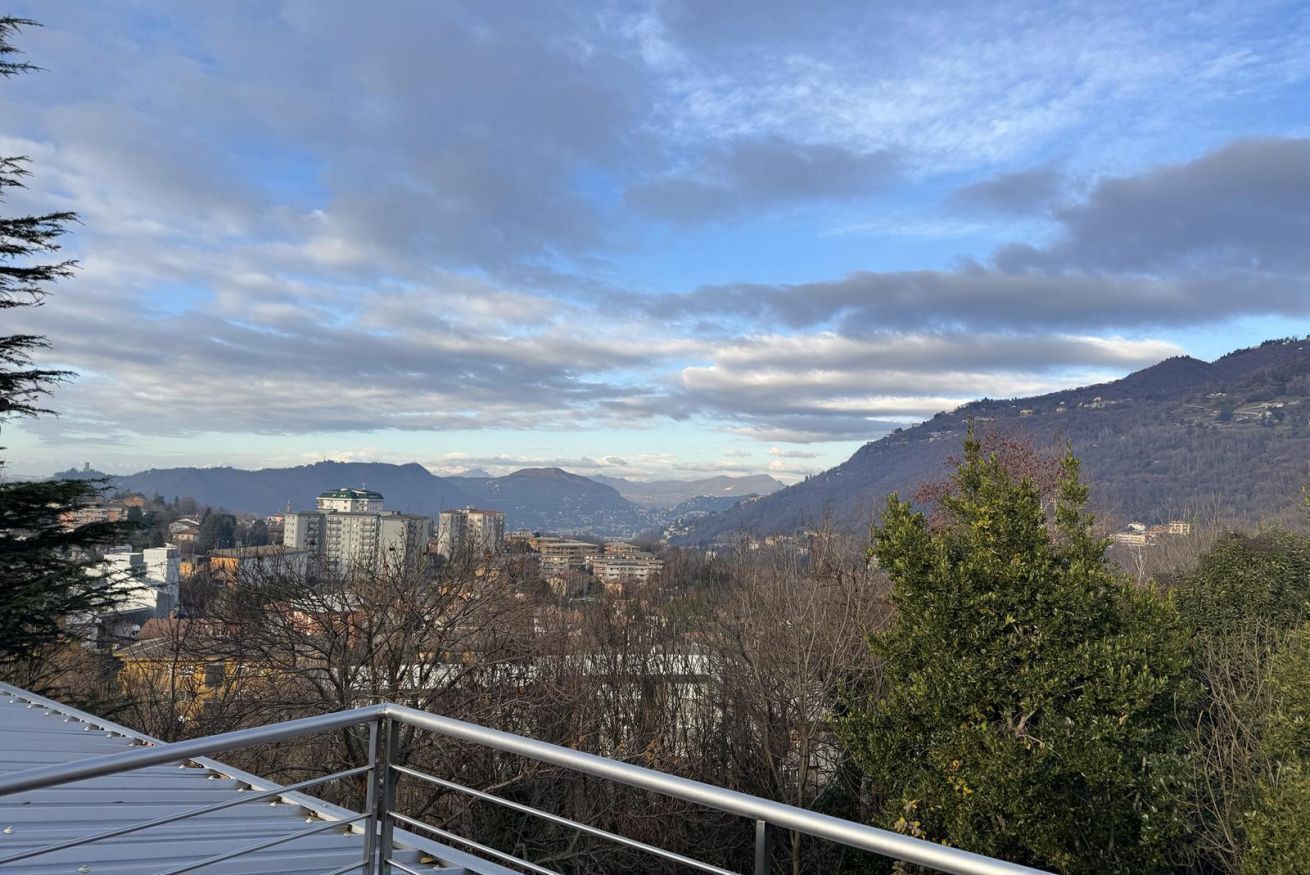 Un paisaje urbano con montañas al fondo, cielo despejado y una barandilla de metal en primer plano.