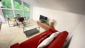 Ein modernes Wohnzimmer mit einem roten Sofa, einem Glastisch und einem Fernseher auf einem Holzboden. Es gibt einen Esstisch mit Stühlen und einen Blick ins Grüne durch große Fenster.