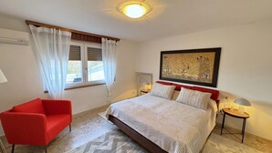 Ein Schlafzimmer mit einem Bett, einem roten Sessel, einem Fenster mit Vorhängen, einem Nachttisch mit einer Lampe und einem gerahmten Bild an der Wand.