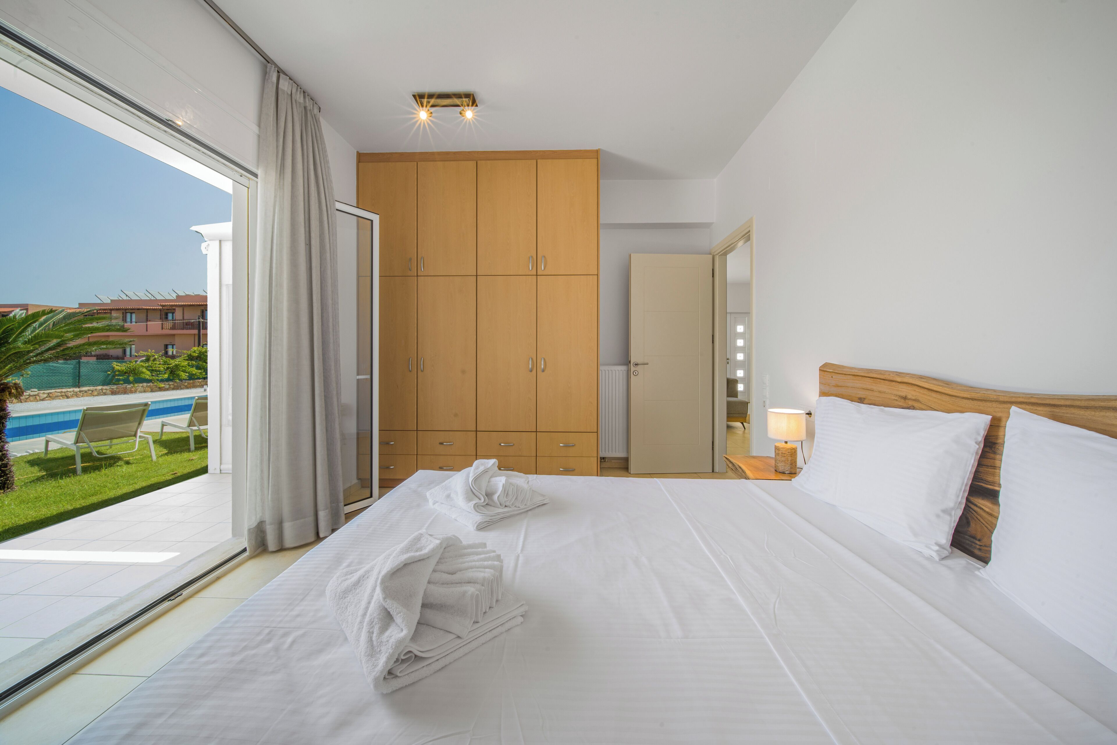 A hotel room with a large bed, a wooden wardrobe, a bathroom, and a view of a pool and greenery.