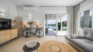A modern living room with a sofa, a television, a dining table with chairs, and a view of a pool through the sliding glass doors. - Villa Yiayia: Relaxing by the sea (Heated pool) (Sisi)