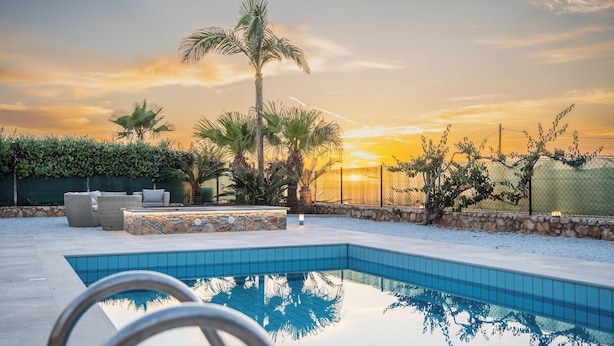 A swimming pool with a stone border, surrounded by a fence, palm trees, and a sunset in the background. - Villa Yiayia: Relaxing by the sea (Heated pool) (Sisi)