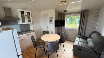 A modern kitchen with a refrigerator, stove, and cabinets. There is a dining table with four chairs and a sofa. A TV is mounted on the wall.