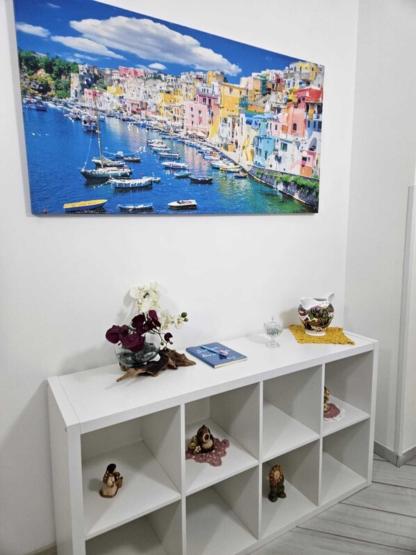 Una consolle bianca con un quadro di una località costiera, un vaso con fiori, un libro e oggetti decorativi.