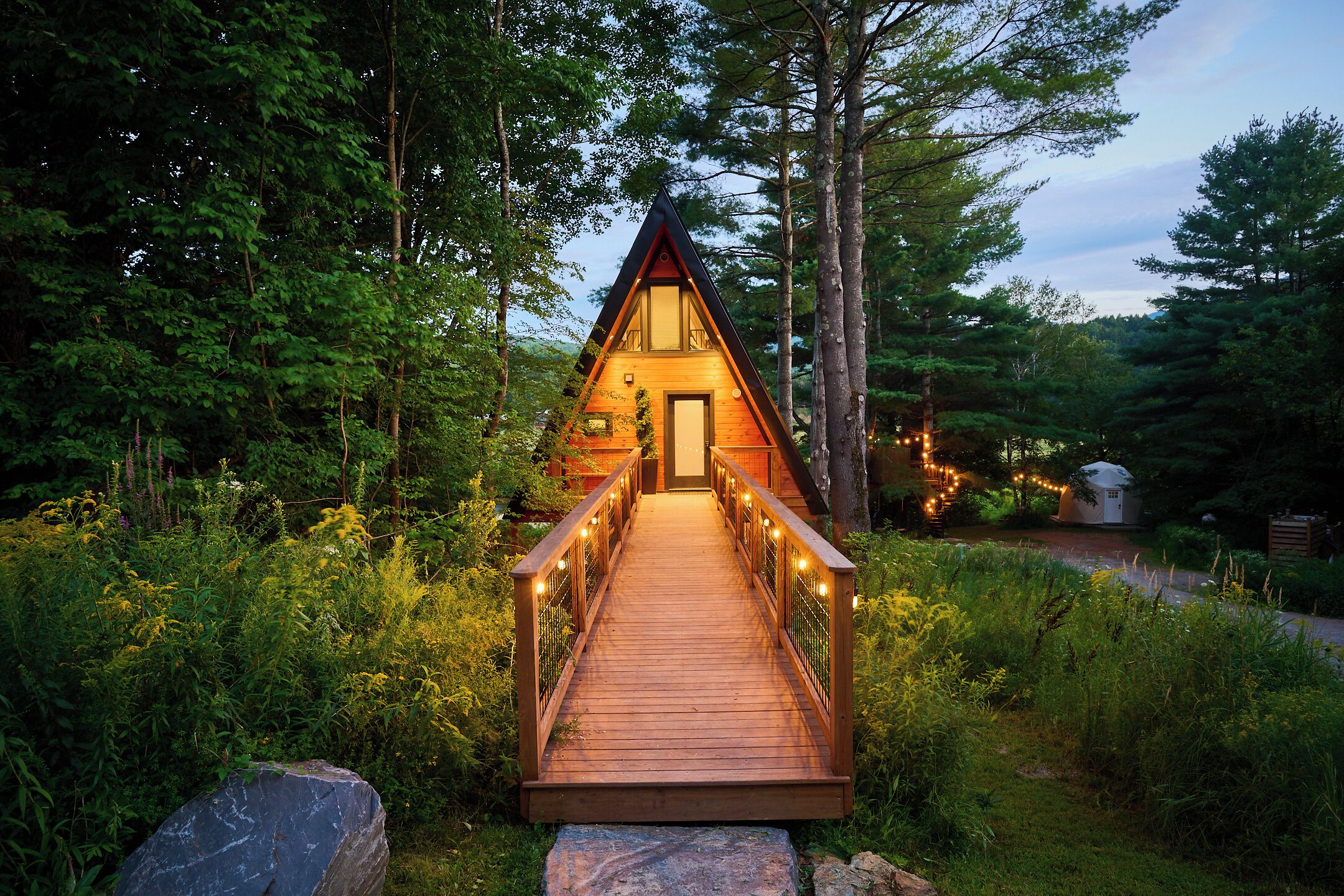 A wooden cabin with a triangular roof, a wooden deck leading to the entrance, surrounded by dense greenery.