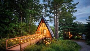 A wooden cabin with a sloped roof, a wooden deck with string lights, surrounded by dense forest.
