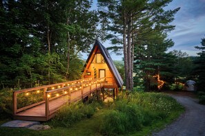A wooden cabin with a sloped roof, a wooden deck with string lights, surrounded by dense forest. - Designer A-Frame Treehouse with hot tub on river (Johnson)