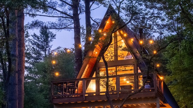 A cabin with a wooden deck and hanging chairs, surrounded by trees. - Designer A-Frame Treehouse with hot tub on river (Johnson)