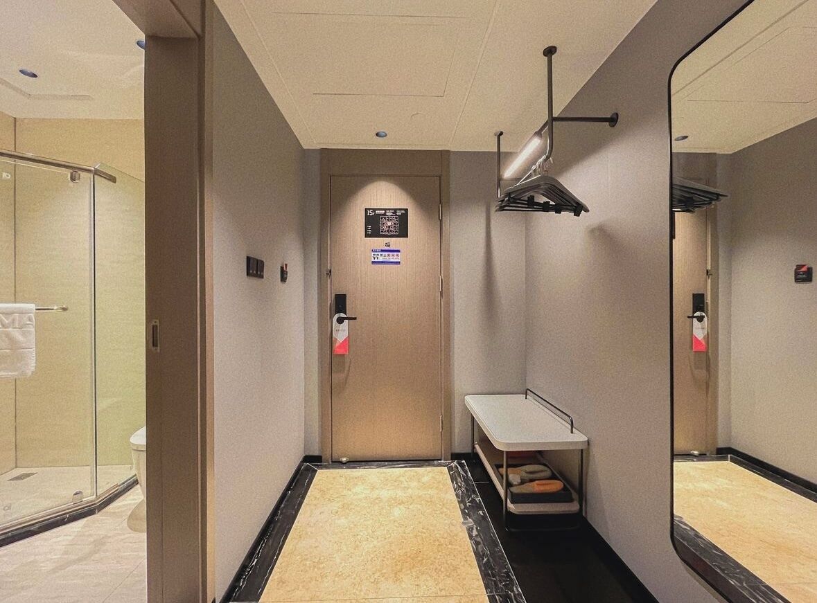 Un couloir d’hôtel avec une douche en verre, une porte dotée d’un code QR, un miroir et une petite table avec des serviettes.