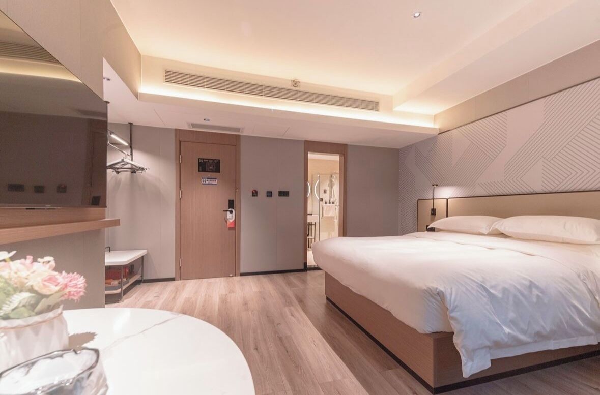 A modern hotel room with a large bed, a bedside table, a mirror, and a small seating area with a vase of flowers.