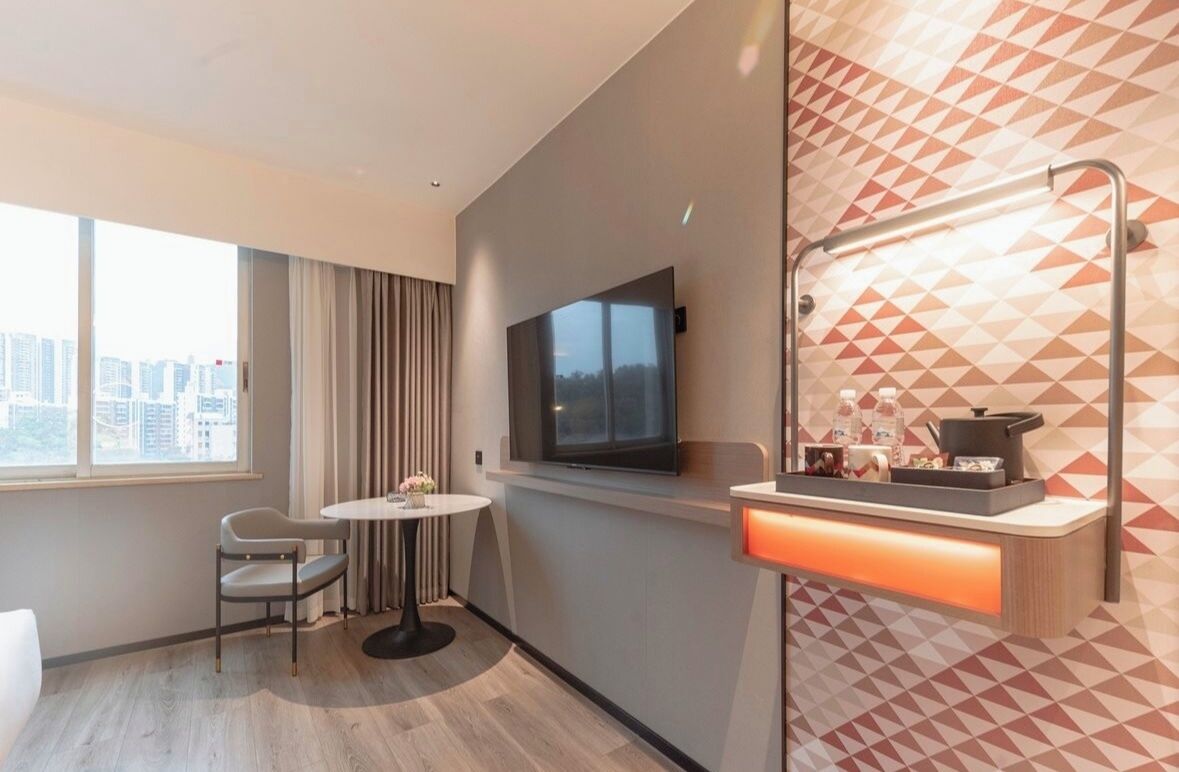 Ein modernes Hotelzimmer mit großem Fenster, Flachbildfernseher, rundem Tisch und einer integrierten Küchenzeile mit Mikrowelle und Wasserkocher.