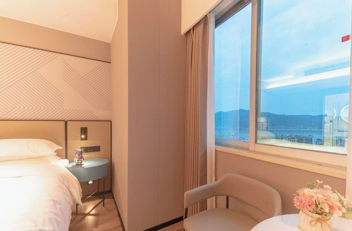 A hotel room with a bed, a small table with a vase of flowers, a chair, a nightstand with a lamp, and a window with a city view.