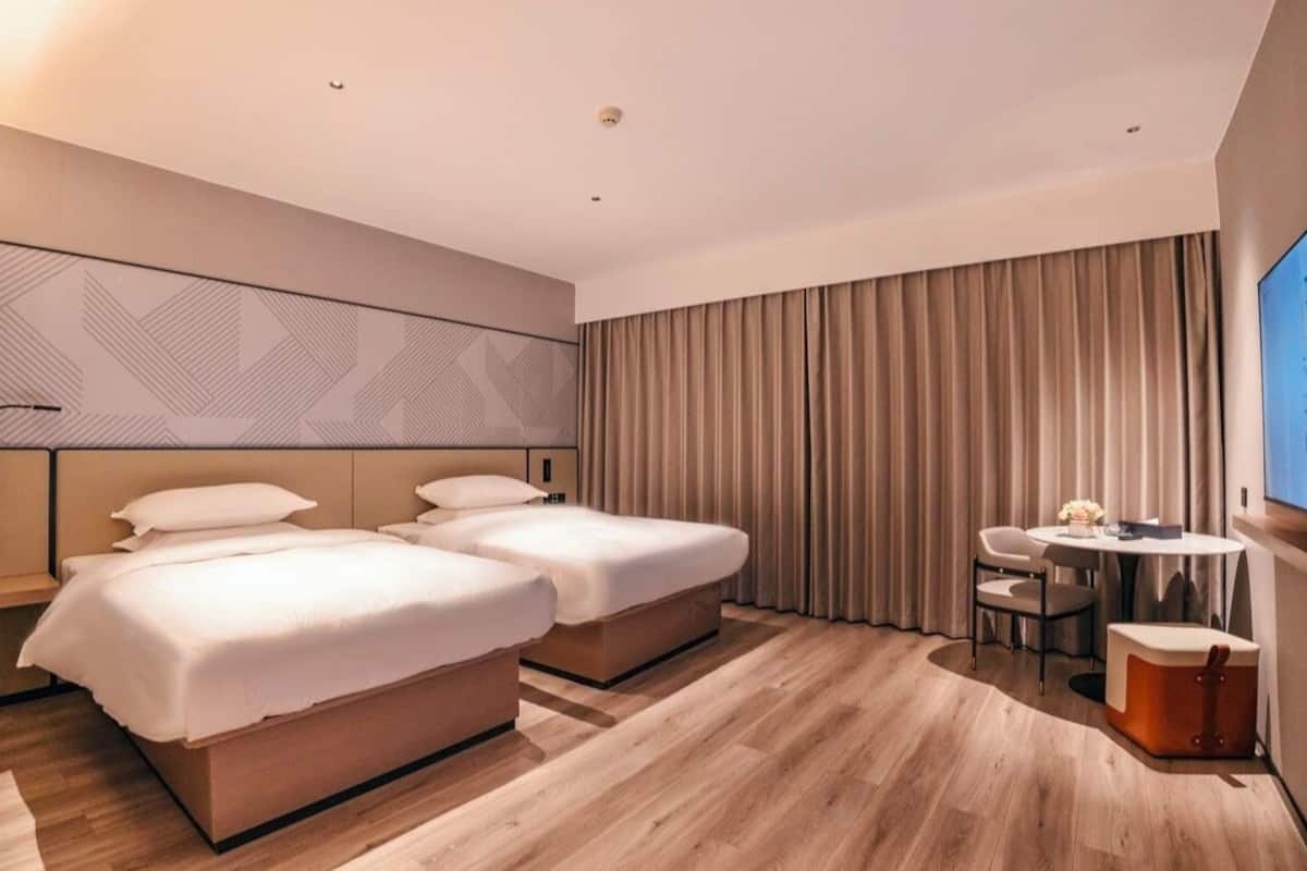 Ett modernt hotellrum med två sängar, ett litet bord med stolar, en TV och ett stort fönster med gardiner.