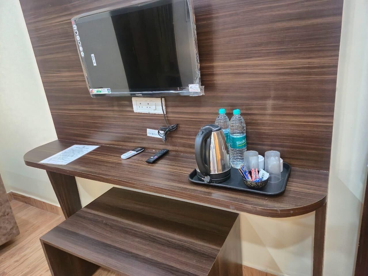 A wooden desk with a flat-screen TV, a kettle, a tray with bottles and glasses, and a remote control.