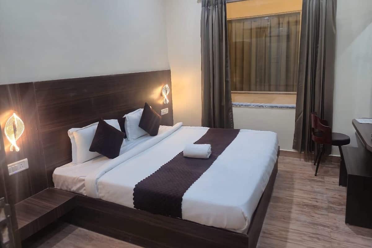 Cama bem arrumada com travesseiros, mesa de cabeceira, escrivaninha e janela com cortinas.