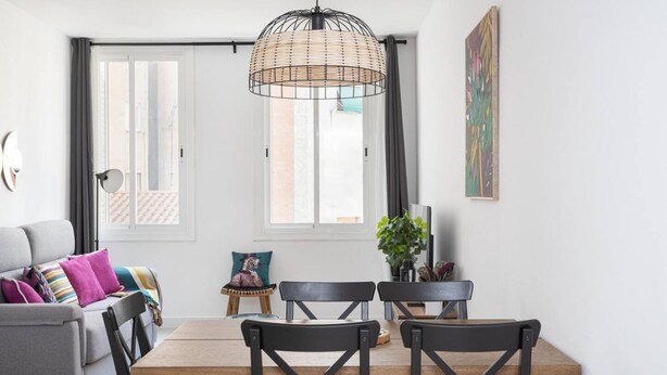 Ett modernt vardagsrum med en grå soffa, ett matbord i trä och svarta stolar. Det finns ett stort fönster med gardiner, en hängande lampa av rotting och en krukväxt.